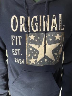 Mörkblå hoodie med stjärntryck - Snygg mörkblå hoodie från Original Fit med stor vit stjärna och texten 'ORIGINAL FIT EST. 2024' på bröstet. Hoodien har huva med dragsko och en klassisk magficka framtill. Perfekt för en avslappnad streetwear-look.