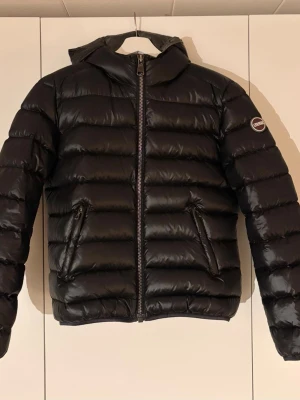 Svart dunjacka från Colmar - Svart pufferjacka (silverfoder) från Colmar med huva och dragkedja framtill. Jackan har två snedställda fickor med dragkedja och ett Colmar-märke på ärmen. Perfekt för kalla dagar och har en klassisk, stilren look. Storlek 44 enligt lapp, skulle säga XS eller mindre S. 