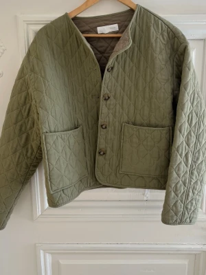 Olivgrön quiltad cardiganjacka - Quiltad cardiganjacka i olivgrönt tyg med raka ärmar och två stora framfickor. Jackan har knäppning med fyra knappar och ett mönster av sydda rutor över hela plagget. Avslappnad passform och rundad halsringning. Perfekt för lager på lager.