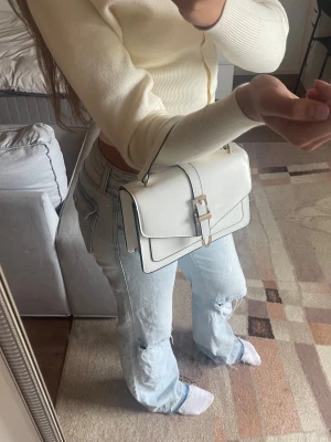Vit liten väska - Snygg vit handväska i slätt konstläder med guldfärgat spänne framtill. Väskan har både ett kort handtag och en avtagbar axelrem, vilket gör den flexibel att bära. Stilren design med raka linjer och svarta kanter vilket gör det lätt att matcha den till både svart och vitt.