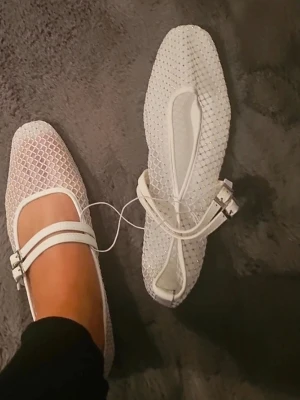 Vita mesh ballerinaskor med spännen - Säljer ett par vita ballerinaskor i mesh med dubbla remmar och spännen. Skorna har fyrkantig tå och transparent nätmaterial som ger en trendig look. Perfekta för dig som gillar stilrena och moderna detaljer.