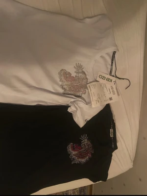 Kenzo t-shirt med drakbroderi - Två snygga Kenzo t-shirts, en vit och en svart, båda med ett detaljerat broderi av en drake på bröstet. Klassisk passform och rund halsringning. Perfekta för dig som vill ha något unikt och trendigt i garderoben.