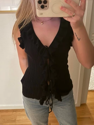 Svart trendig topp Zara - Trendig svart topp med volangdetaljer längs urringningen och axlarna. Toppen har knytband framtill och är gjord i ett ribbat, stretchigt material som sitter snyggt på kroppen. Perfekt för dig som gillar en feminin och cool stil.