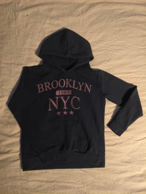 Mörkblå hoodie med tryck - Mörkblå hoodie med trycket 'BROOKLYN 1989 NYC' i lila på bröstet. Klassisk känguruficka framtill och ribbade muddar vid ärmslut och nederkant. Perfekt för en avslappnad streetwear-look. Storlek 146-152 men passar även till de som har storlek 134-140. 