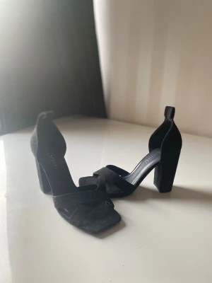 Svarta klackar med bred klack - Stilrena svarta klackar skor med bred klack och öppen tå. Skorna har en trendig fyrkantig tå och remmar i mockaimitation som korsar över foten. Perfekta för dig som vill ha både höjd och stabilitet. Passar till många olika looks.