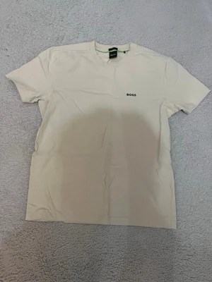 Vit t-shirt från BOSS - Snygg vit t-shirt från BOSS med minimalistisk logga på bröstet. Klassisk rund halsringning och korta ärmar. Perfekt basplagg i mjukt bomullsmaterial som passar till allt.