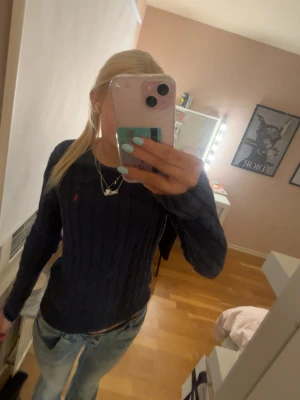 Mörkblå stickad tröja Polo Ralph Lauren - Mörkblå kabelstickad tröja från Polo Ralph Lauren i mjuk Pima bomull. Klassisk passform med rund halsringning och lång ärm. Liten röd logga broderad på bröstet. Perfekt till jeans för en clean och stilren look.