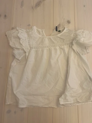 Vit blus med volangdetaljer - Söt vit blus med korta ärmar och volangdetaljer längs axlarna. Blusen har spetsinslag framtill och en lös, luftig passform. Perfekt till jeans eller kjol för en romantisk look. Tillverkad i ett lätt bomullsliknande material.