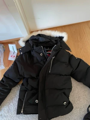Svart dunjacka Moose Knuckles - Snygg svart dunjacka från Moose Knuckles med avtagbar huva och vit fuskpäls. Jackan har dragkedja, tryckknappar och flera fickor med dragkedja. Perfekt för kalla vinterdagar och riktigt varm.