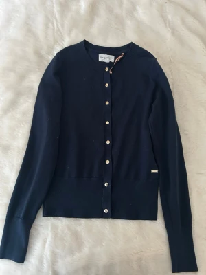 Mörkblå kofta från Holly & Whyte - Klassisk mörkblå kofta från Holly & Whyte by Lindex med rund halsringning och guld knappar framtill. Tunnstickad modell med ribbade muddar och diskret logodetalj vid nederkanten. Perfekt att slänga över en topp för en stilren look.