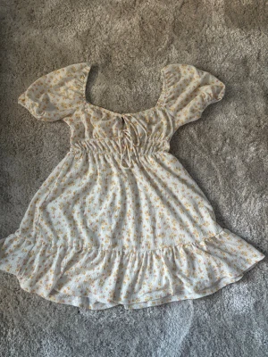 Blommig kortklänning Gina Tricot - Supersöt kortklänning från Gina Tricot i vitt med små gula och orange blommor. Klänningen har puffärmar, snörning framtill och volangkant nedtill. Perfekt för sommarens alla tillfällen!