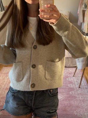 Beige kofta med fickor och knappar - Mysig beige kofta med v-ringning och stora knappar framtill. Två praktiska fickor på magen och ribbade muddar vid ärmslut och nederkant. Perfekt att slänga över en t-shirt för en avslappnad look.