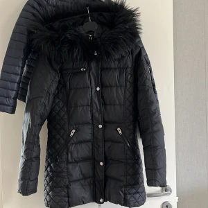 Svart jacka äkta - Snygg svart dunjacka från Rockandblue med avtagbar huva och fuskpäls. Jackan har dragkedjor vid fickorna, quiltade detaljer och Rockandblue-logga på ärmen. Perfekt för kalla vinterdagar och ger en trendig look.