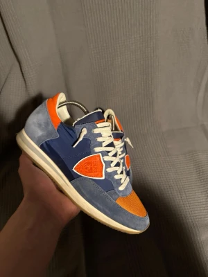 Philippe Model - Snygga sneakers från Philippe Model i blå mocka och textil med orange detaljer och vita skosnören. Skorna har en klassisk låg siluett och en vit sula som ger en retrovibe. Perfekta för dig som vill sticka ut med färg och stil.