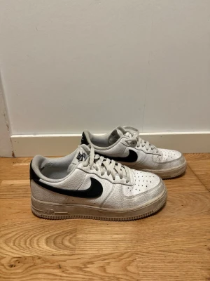 Nike Air Force 1 vita med svart swoosh - Klassiska Nike Air Force 1 sneakers i vitt läder med svart swoosh på sidorna. Lågt skaft och perforerad tå för extra ventilation. Tjock sula med Air-dämpning och Nike-logga på plösen. Perfekta för en streetwear-look.