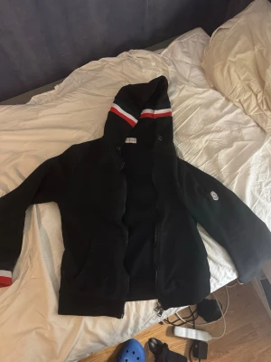 Svart hoodie med rödvit detalj - Säljer en svart hoodie med dragkedja och huva. Huvan har snygga rödvita ränder och ärmarna har matchande ränder vid mudden. På vänster ärm finns en liten rund logga. Hoodien har fickor framtill och är i mjukt material.