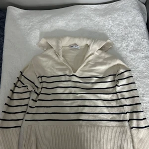 Randig stickad tröja från Zara - Säljer en snygg stickad tröja från Zara i storlek XS. Tröjan är krämvit med marinblå ränder, har en bred krage med v-ringning och långa ärmar. Ribbad nederkant och muddar ger en chill vibe. Perfekt för dig som gillar klassisk stil med en twist. Den är oanvänd.