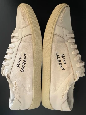 Saint Laurent vita sneakers - Säljer ett par vita Saint Laurent sneakers med handskriven logga på sidan. Skorna har råa, slitna kanter och detaljer i canvas och skinn. Klassisk rund tå, platt sula och snörning framtill. Perfekt för dig som gillar en edgy och avslappnad stil. Bra skick med mindre defekter som lite uppfläkt innuti, sulan har släppt pyttelite vid några få ställen och ett av slitdetaljerna har nått ner en bit extra men inget som direkt påverkar eller syns.🙌
