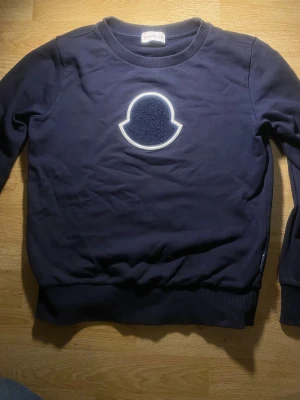 Mörkblå sweatshirt från Moncler - Mörkblå sweatshirt från Moncler med stort vitt och blått logomärke på bröstet. Tröjan har rund hals, ribbade muddar vid ärmslut och nederkant samt en mjuk bomullskänsla. Perfekt för dig som gillar stilrena och exklusiva plagg.