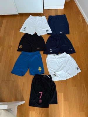 Fotbollsshorts barn - Säljer ett gäng fotbollsshorts från olika lag och märken som Nike, Adidas. Färgerna är svart, vit, blå och marinblå. Shorts med klubbmärken som Real Madrid, Inter, Tottenham, PSG och svenska landslaget. Perfekta för träning eller match! Storlekarna varierar, men de är i barn. Tveka inte på att höra av dig vid funderingar!  50kr styck, chans för billigare stickprovs vid köp av flera.