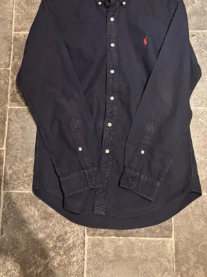 Mörkblå skjorta från Ralph Lauren - Klassisk mörkblå skjorta från Ralph Lauren med röd broderad logga på bröstet. Skjortan har långa ärmar, knappar framtill och button-down krage. Tillverkad i mjuk bomull som känns skön mot huden. Perfekt för en clean och stilren look.