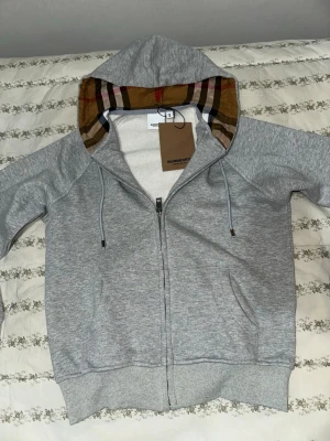 Burberry kofta - Snygg grå hoodie med dragkedja från Burberry. Klassisk huva med det ikoniska rutmönstret på insidan, dragsnören och två fickor framtill. Tillverkad i mjukt bomullsmaterial för en bekväm känsla. Den är liten i storlek så skulle säga den passar S mer.