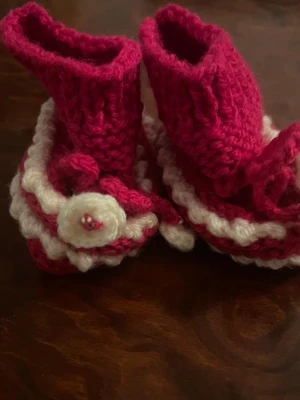 Handstickade röda och vita tofflor - Säljer ett par handstickade tofflor i rött och vitt med dekorativ blomdetalj framtill. Tofflorna har högt skaft och är gjorda i mjukt garn, perfekta för att hålla fötterna varma inomhus. Passar dig som gillar unika och mysiga accessoarer.