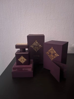 Initio Side Effect Eau de Parfum Parfym - Lyxig parfym från Initio Parfums Privés i en elegant lila flaska med guldfärgade detaljer och dekorativt mönster. Flaskan har en fyrkantig form och kommer i matchande lila förpackning med gulddetaljer. Perfekt för dig som gillar exklusiva dofter och snygg design.