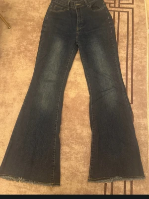 Mörkblå utsvängda jeans - Säljer ett par mörkblå jeans med utsvängda ben och rå kant längst ner. Klassisk femficksmodell med hög midja och slitningar på låren för en cool vintagekänsla. Perfekta för dig som gillar retrostil och vill sticka ut lite extra.