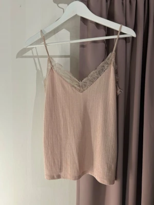 Ljusbeige ribbad linnetopp med spets - Säljer en ljusbeige ribbad linnetopp med tunna axelband och vacker spetsdetalj längs v-ringningen. Toppen har en mjuk och feminin look, perfekt för att styla lager på lager. Materialet känns lätt och luftigt. Passar även S/M