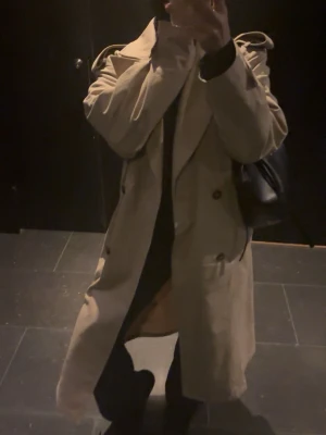 Beige trenchcoat med bred krage - Stilren beige trenchcoat med dubbelknäppning och bred krage. Jackan har klassiska axelklaffar och ett avslappnat, oversized snitt. Tillverkad i ett mjukt tyg som ger en snygg drapering. Perfekt för dig som gillar tidlös och trendig stil.