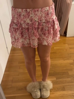 Rosa volangkjol med blommönster - Supersöt kort kjol i vitt och rosa med blommigt mönster och volangdetaljer. Kjolen har bred smock i midjan för skön passform och är gjord i ett lätt, luftigt material som passar perfekt till varma dagar.