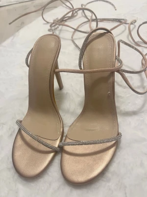 Nude pumps med strass och snörning - Snygga pumps i nude färg med tunna remmar dekorerade med strass och lång snörning runt ankeln. Skorna har hög klack och öppen tå.  Materialet ser ut att vara satinliknande . Aldrig använt de