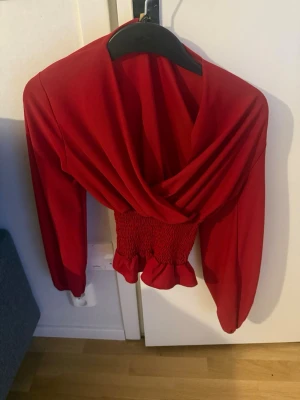 Röd omlottopp med smockad midja - Trendig röd omlottopp med lång ärm och smockad, volangkantad midja. Toppen har en djup v-ringning och är croppad, vilket ger en snygg siluett. Perfekt för dig som vill sticka ut med färg och form.