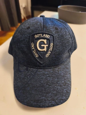 Mörkblå Gotland keps med brodyr - Snygg mörkblå keps med broderad Gotland-logga och texten 'Gotland' runt ett stort G på framsidan. Kepsen har böjd skärm och justerbar passform. Materialet är mjukt och har en melerad look som ger en modern vibe.
