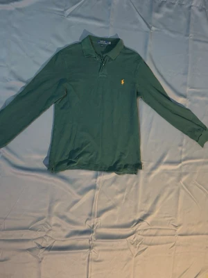 Grön långärmad piké från Polo Ralph Lauren - Klassisk grön långärmad pikétröja från Polo Ralph Lauren med gul broderad logga på bröstet. Slim fit-modell med krage och knappar vid halsen. Perfekt till jeans eller chinos för en clean och stilren look.