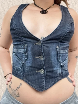 Mörkblå jeansväst med knappar - Snygg mörkblå jeansväst  med tre knappar framtill och två fickor med lock. Västen har en figurnära passform och coola sömdetaljer. Perfekt att styla över en t-shirt eller klänning för en trendig look.