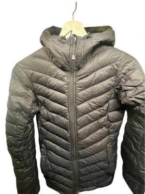 Svart dunjacka Peak Performance - Snygg svart dunjacka från Peak Performance med huva och dragkedja framtill. Jackan har ett stilrent, quiltat mönster och diskret logga på bröstet. Perfekt för kalla dagar och enkel att matcha med olika outfits.