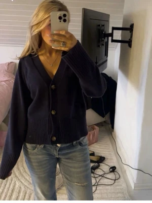 Mörkblå stickad kofta med knappar - Säljer en mörkblå stickad kofta med stora knappar framtill och v-ringad hals. Koftan har en avslappnad passform, ribbade muddar och är lite croppad i modellen. Perfekt att slänga över en t-shirt eller linne.