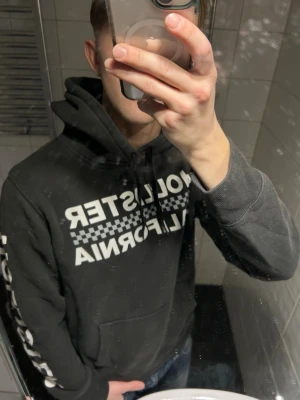 Svart Hollister hoodie med tryck - Svart hoodie från Hollister med vit text och schackrutigt tryck på bröstet samt text på ärmen. Klassisk känguruficka framtill och justerbar huva med snören. Perfekt för en avslappnad och cool stil.