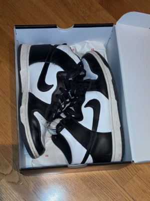 Nike Dunk High Panda sneakers - Svarta och vita Nike Dunk High sneakers med klassisk svart swoosh på sidan. Skorna har högt skaft, vita paneler och svarta detaljer i läder samt svart snörning. Sulan är vit med svart yttersula. Perfekt för dig som gillar streetwear och tidlös stil.