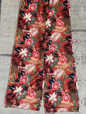 Blommiga byxor från Zara - Färgglada vida kostymbyxor från Zara med tropiskt blommönster i rött, rosa, vitt och grönt. Vida ben och hög midja med dragkedja och knapp framtill. Perfekta för dig som gillar statement-plagg och vill sticka ut med en unik stil. OBS - sömmen längst ner på ena benet har gått upp och behöver sys till, inte svårt men det är lite längre än det andra benet som det är nu. 