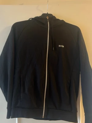 Svart hoodie från Hugo Boss - Snygg svart hoodie från Hugo Boss med dragkedja framtill och diskret BOSS-logga på bröstet. Klassisk design med huva och dragsko, samt fickor på sidorna. Perfekt för en clean och stilren look.