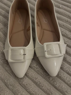 Vita ballerinaskor med stor spänne - Snygga vita ballerinaskor från New Look med spetsig tå och en bred rem med stor fyrkantig spänne framtill. Skorna har en glansig finish och platt sula, vilket ger en stilren och modern look. Perfekta för dig som gillar minimalistisk design med en twist.
