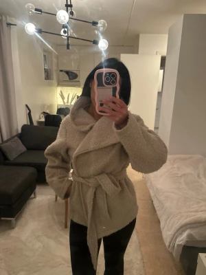 Beige teddyjacka med knytskärp - Säljer en beige teddyjacka med oversized krage och knytskärp i midjan. Jackan har en mjuk, fluffig yta och långa ärmar. Perfekt för dig som vill ha en mysig och trendig look under kalla dagar.