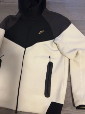 Nike svartvit vindjacka med dragkedja - Nike vindjacka i svart och vitt med hög krage och hel dragkedja framtill. Jackan har två fickor med dragkedja och Nike-logga på bröstet. Tillverkad i mjukt syntetmaterial och har långärmad design. Perfekt för en sportig och clean look.