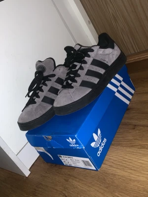 Adidas Campus grå/svarta sneakers - Snygga Adidas Campus sneakers i grå mocka med svarta detaljer och klassiska tre ränder på sidan. Skorna har svart sula, svarta skosnören och Adidas-logga på plösen. Perfekta för dig som gillar stilrena och tidlösa sneakers. Köpta för 1319kr