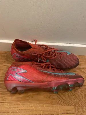 Röda Nike Mercurial elite  - Säljer ett par röda Nike Mercurial fotbollsskor med turkosa detaljer och snörning. Skorna har en låg profil och är tillverkade i syntetmaterial för bra grepp och kontroll på planen. Perfekta för dig som vill ha lätta och snabba skor till match eller träning.