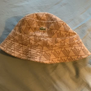 Beige bucket hat från Lacoste - Säljer en beige bucket hat från Lacoste med geometriskt mönster i brunt och det klassiska gröna krokodilmärket framtill. Hatten är i mjukt tyg och har en avslappnad passform som ger en cool och trendig vibe.