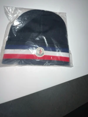 Mörkblå Moncler mössa med ränder - Snygg mörkblå mössa från Moncler med klassiska ränder i blått, vitt och rött samt Moncler-logga framtill. Perfekt accessoar för att hålla värmen och samtidigt se trendig ut. Tillverkad i mjukt stickat material.